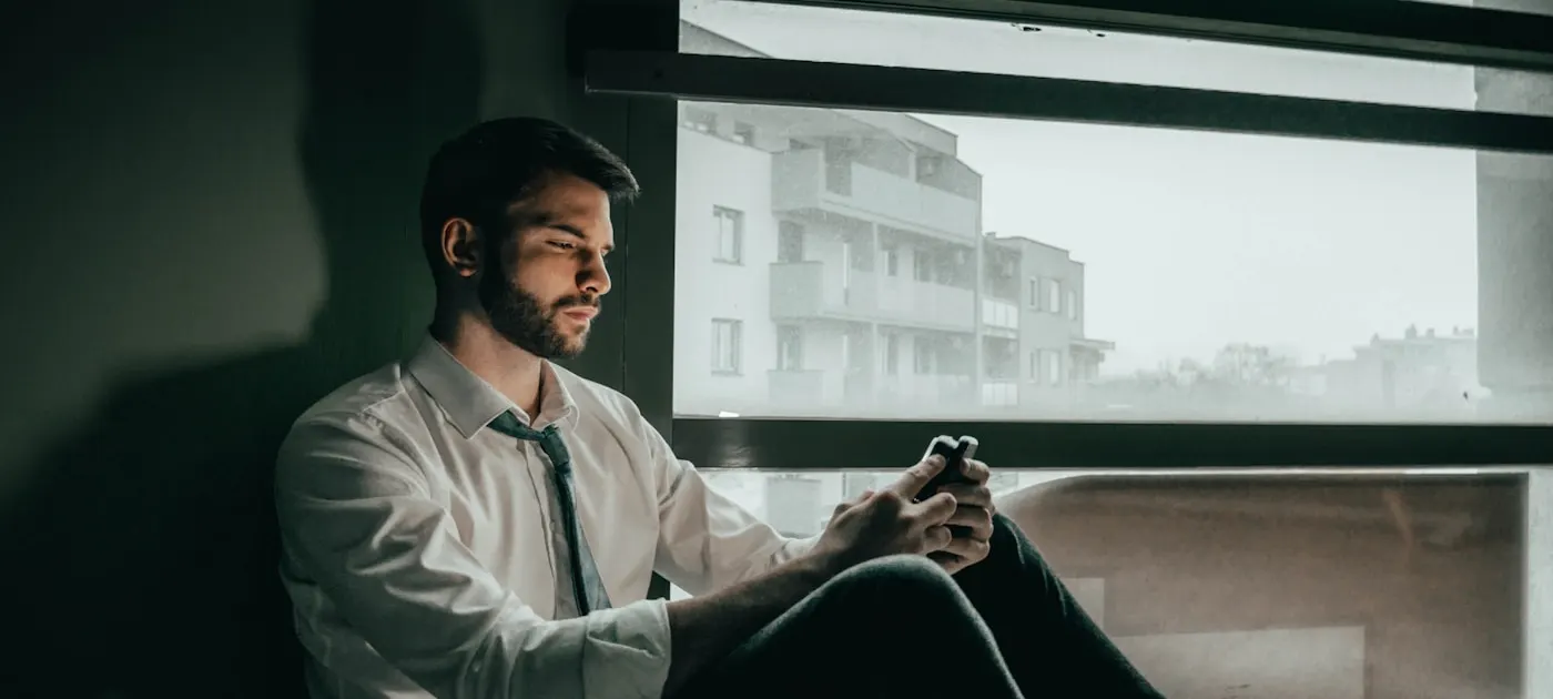 a man sitting on a window sill looking at his cell phone