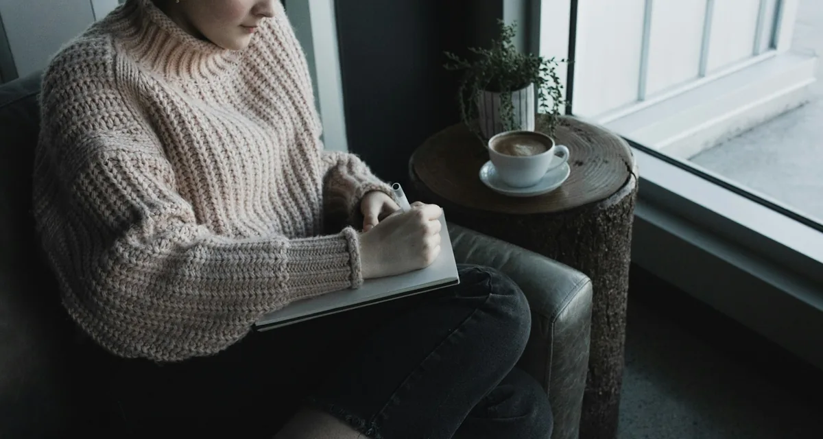 A person writing in a notebook beside a cup of coffee in the light of a window